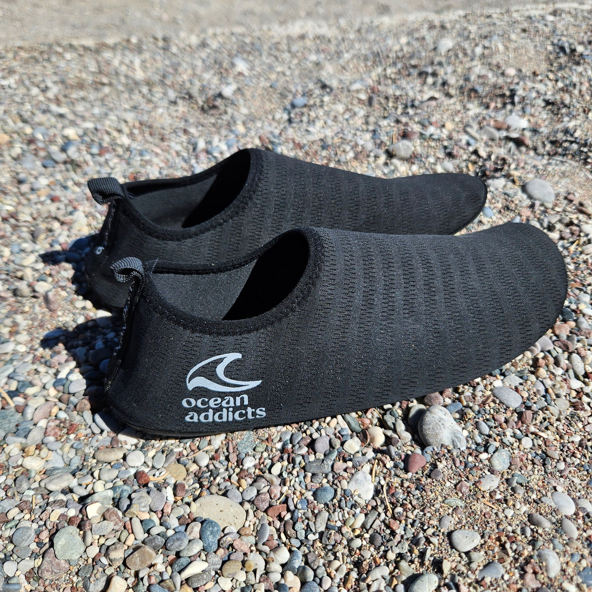 Sorte badesko på stenstrand med hvidt Ocean Addicts-logo