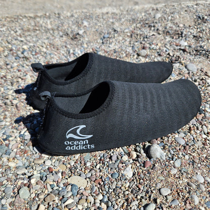 Sorte badesko på stenstrand med hvidt Ocean Addicts-logo