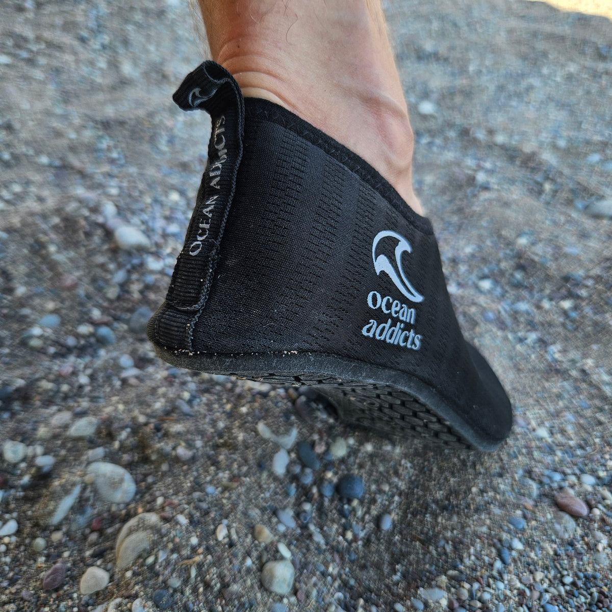 Sort badesko på fod ved stenstrand med Ocean Addicts-logo