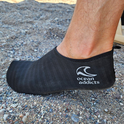 Sort badesko på fod på stenet strand med Ocean Addicts-logo