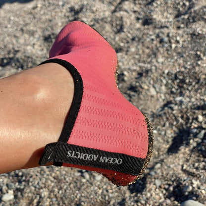 Koralfarvet badesko med sort kant på fod ved stenet strand